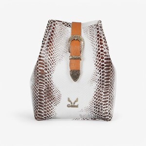 Op de afbeelding: Een witte en bruine rugzak met slangenpatroon, met een bruine leren riem en zilveren gesp. De tas heeft een unieke vorm en draagt de merknaam "Genni" in zilver.
