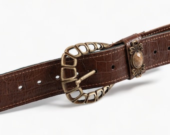 May include: A brown leather belt with a decorative, ornate, gold-toned buckle. The buckle features a raised, textured design and a small, clear stone.