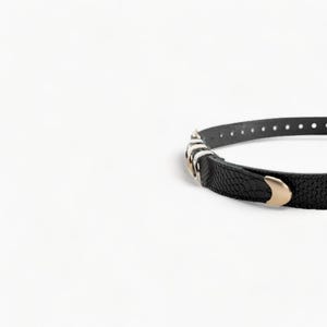 May include: A black leather belt with a gold buckle and silver accents. The belt has a textured surface and is designed to be worn around the waist.