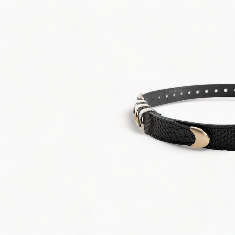 May include: A black leather belt with a gold buckle and silver accents. The belt has a textured surface and is designed to be worn around the waist.