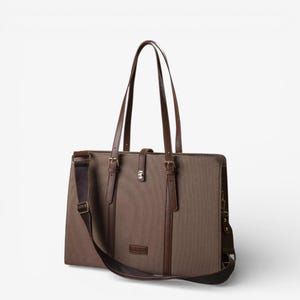 Può includere: Una borsa tote in tela marrone con dettagli in pelle. La borsa è dotata di tracolla e due manici superiori. Presenta cinturini in pelle con fibbie e una piccola toppa rettangolare in pelle. La borsa è progettata per trasportare oggetti.