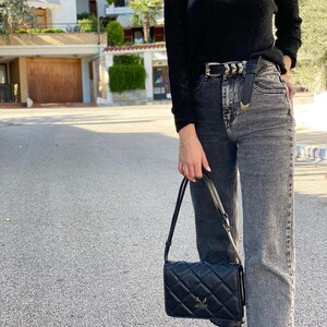May include: A woman wearing a black sweater, a black belt with a silver buckle, and black and grey jeans. She is carrying a black quilted shoulder bag with a gold logo.