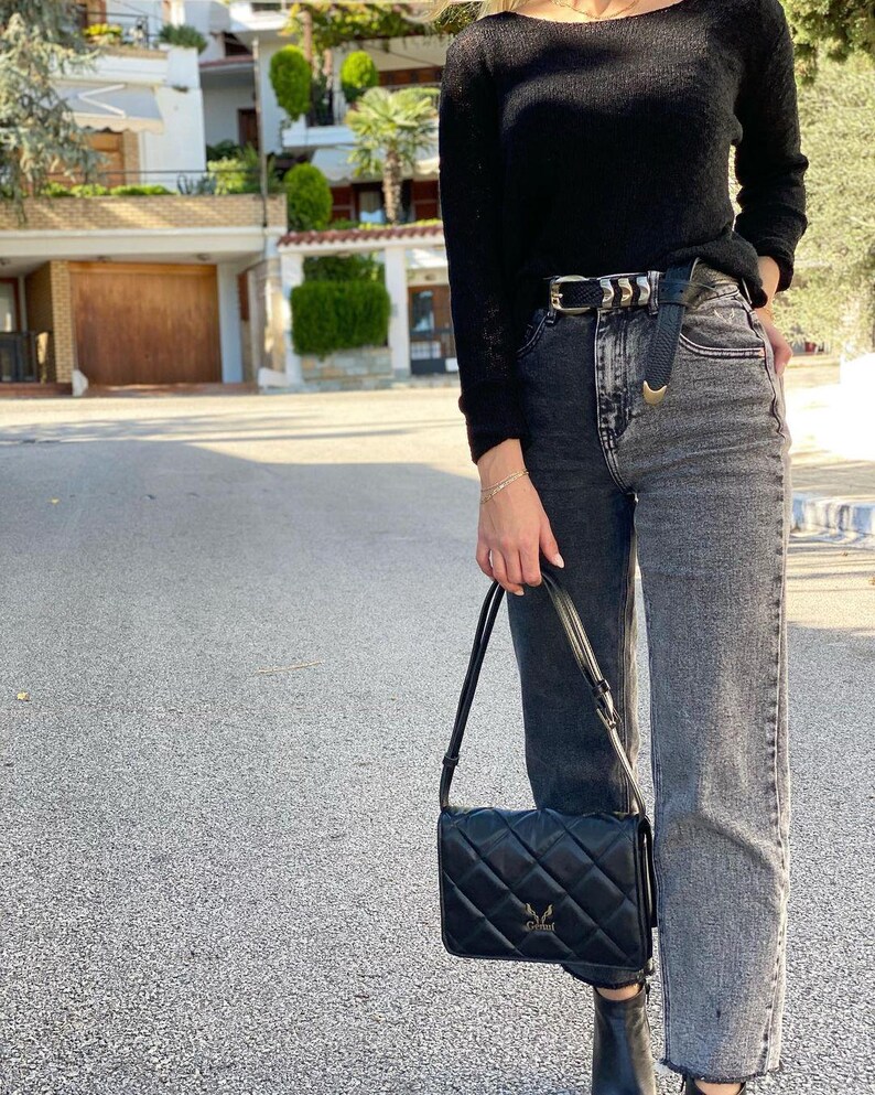 May include: A woman wearing a black sweater, a black belt with a silver buckle, and black and grey jeans. She is carrying a black quilted shoulder bag with a gold logo.