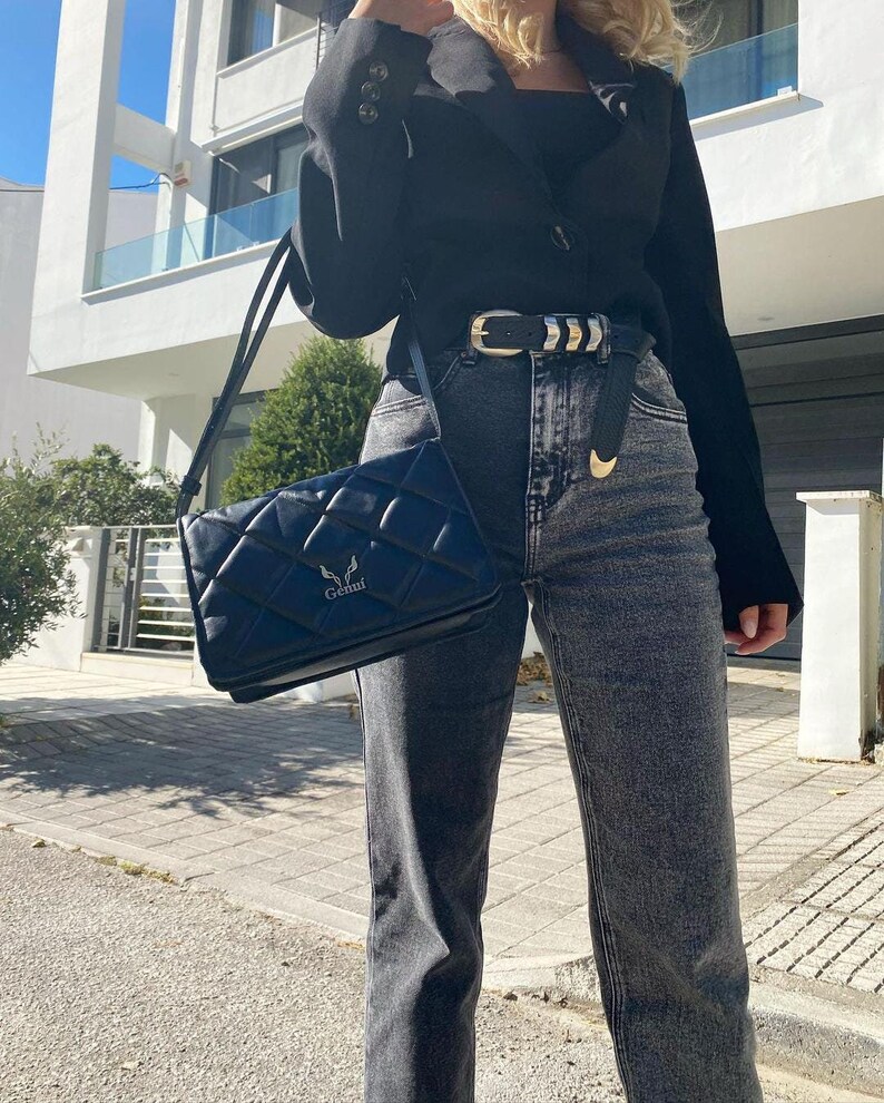 May include: A woman wearing a black blazer, black top, and dark wash jeans with a silver belt. She is carrying a black quilted shoulder bag with a silver logo that says "Genuf."