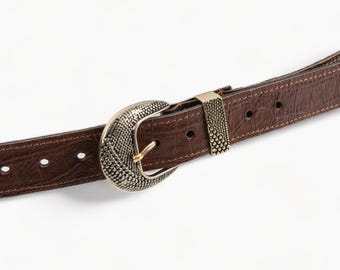 May include: A brown leather belt with a decorative, ornate, gold-toned buckle. The buckle is oval-shaped and has a textured design.
