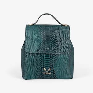 Puede incluir: Una mochila de cuero verde con estampado de piel de serpiente y un logotipo dorado que dice "Gemut".