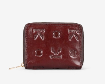 May include: A burgundy leather wallet with a zip closure and embossed designs. The wallet has a small gold zip pull.