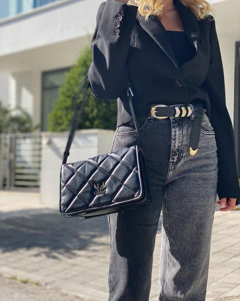 May include: A black quilted shoulder bag with a gold metal logo that says "Genny". The bag is worn over a black blazer and dark wash denim jeans with a gold and black belt.