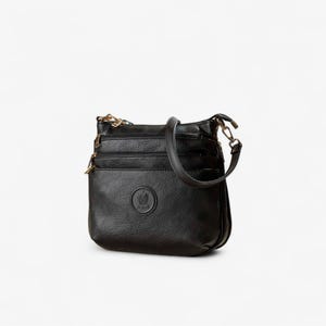 Op de afbeelding: Zwarte leren crossbodytas met meerdere vakken met ritssluiting en een schouderriem. De tas heeft goudkleurige hardware en een rond embleem. De tas is ongeveer 25 cm breed en 20 cm hoog.