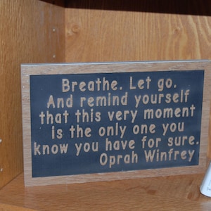 May include: A framed sign with the quote "Breathe. Let go. And remind yourself that this very moment is the only one you know you have for sure. Oprah Winfrey." The sign has a dark blue background and a wooden frame.