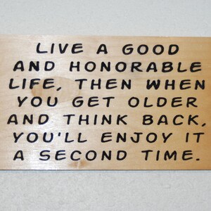 以下が含まれることがあります： 黒い文字で「LIVE A GOOD AND HONORABLE LIFE, THEN WHEN YOU GET OLDER AND THINK BACK, YOU'LL ENJOY IT A SECOND TIME.」と書かれた長方形の明るい色の木製プレート。木の表面には木目と小さな円形のシミが見えます。