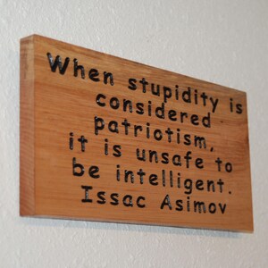 Puede incluir: Placa rectangular de madera con la cita "When stupidity is considered patriotism, it is unsafe to be intelligent. Issac Asimov" en letras negras. La madera tiene un tono cálido y natural.