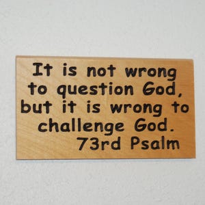 Puede incluir: Un letrero rectangular de madera con el texto "It is not wrong to question God, but it is wrong to challenge God. 73rd Psalm" en letras negras. El letrero es de color marrón claro y está montado sobre una superficie blanca.