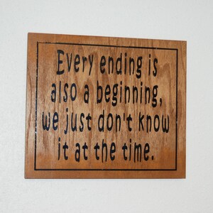 May include: A wooden sign with a black border and the text "Every ending is also a beginning, we just don't know it at the time."