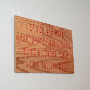 May include: A rectangular wooden plaque with the quote "IF YOU ARE MORE FORTUNATE THAN OTHERS, BUILD A LONGER TABLE, NOT A TALLER FENCE" in red. The plaque is made of light brown wood and is mounted on a white wall.