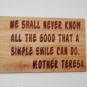 Könnte beinhalten: Helle Holztafel mit dem Zitat "WE SHALL NEVER KNOW ALL THE GOOD THAT A SIMPLE SMILE CAN DO. MOTHER TERESA" in erhabenen, braunen Buchstaben. Die Tafel hat eine helle Holzmaserung.