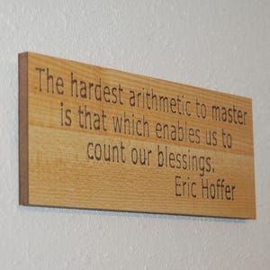 Puede incluir: Un letrero rectangular de madera con la cita "La aritmética más difícil de dominar es la que nos permite contar nuestras bendiciones". El nombre del autor, Eric Hoffer, está debajo de la cita.