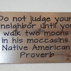 May include: Wooden sign featuring the quote "Do not judge your neighbor until you walk two moons in his moccasins. Native American Proverb" in black text. The sign has a wood grain.
