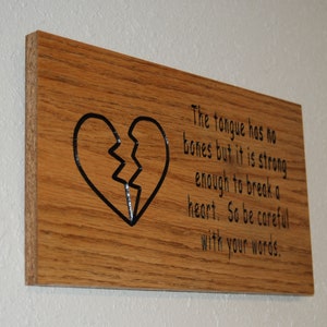 Puede incluir: Un letrero de madera con un corazón roto negro y el texto "La lengua no tiene huesos, pero es lo suficientemente fuerte como para romper un corazón. Así que ten cuidado con tus palabras."