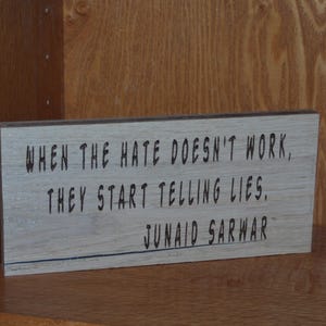 May include: A rectangular wooden sign with the quote "WHEN THE HATE DOESN'T WORK, THEY START TELLING LIES. JUNAID SARWAR" in black lettering. The sign is light brown and sits on a wooden shelf.