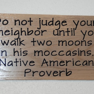May include: A rectangular wooden sign with the quote "Do not judge your neighbor until you walk two moons in his moccasins. Native American Proverb" in black text. The sign is light brown.