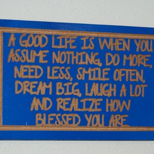 以下が含まれることがあります： 茶色の縁取りのある青い木製の看板に、「A good life is when you assume nothing, do more, need less, smile often, dream big, laugh a lot and realize how blessed you are.」という文字が書かれている。