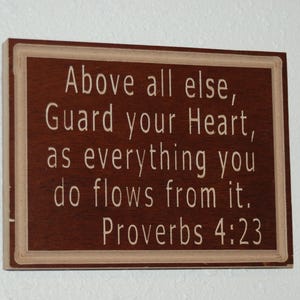 以下が含まれることがあります： 茶色の背景に白い文字が書かれた木製の看板。看板には「Above all else, Guard your Heart, as everything you do flows from it. Proverbs 4:23」と書かれています。