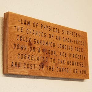 May include: A wooden sign with the text " -LAW OF PHYSICAL SURFACES- THE CHANCES OF AN OPEN-FACED JELLY SANDWICH LANDING FACE DOWN ON A FLOOR, ARE DIRECTLY CORRELATED TO THE NEWNESS AND COST OF THE CARPET OR RUG."