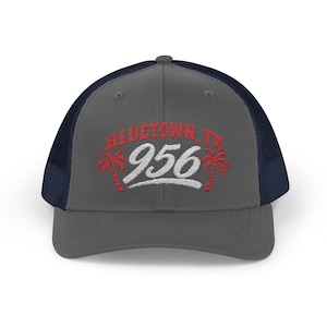 May include: A charcoal gray baseball cap with a navy blue mesh back. The front features embroidered red text reading BLUETOWN, TX above the white number 956, flanked by two red palm tree icons on either side of the central white digits.