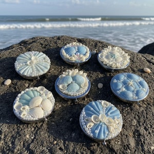 Pode incluir: Sete espelhos compactos decorativos com desenhos de conchas. Cada compacto apresenta um arranjo diferente de conchas brancas e azuis, pérolas e uma borda prateada. Os espelhos são exibidos em uma rocha cinza escura com o oceano ao fundo.