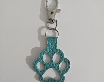 May include: A teal paw print keychain with a silver-colored clasp. The paw print is made of textured material and has a small loop for attachment to the clasp. The background is plain white.