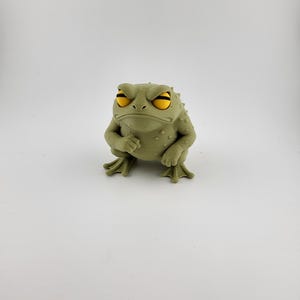 May include: A grumpy-looking, olive-green frog figurine with yellow eyes. The frog has a textured skin, a furrowed brow, and is posed with its fists clenched. The figurine is set against a plain white background.