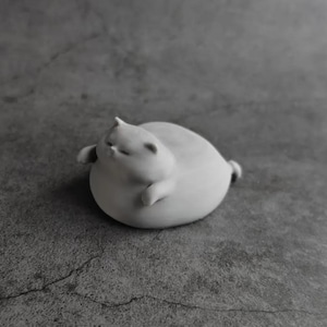 May include: A small, white, ceramic cat figurine in a relaxed pose. The cat is round with its eyes closed, small ears, and tiny paws. The figurine is on a gray surface.