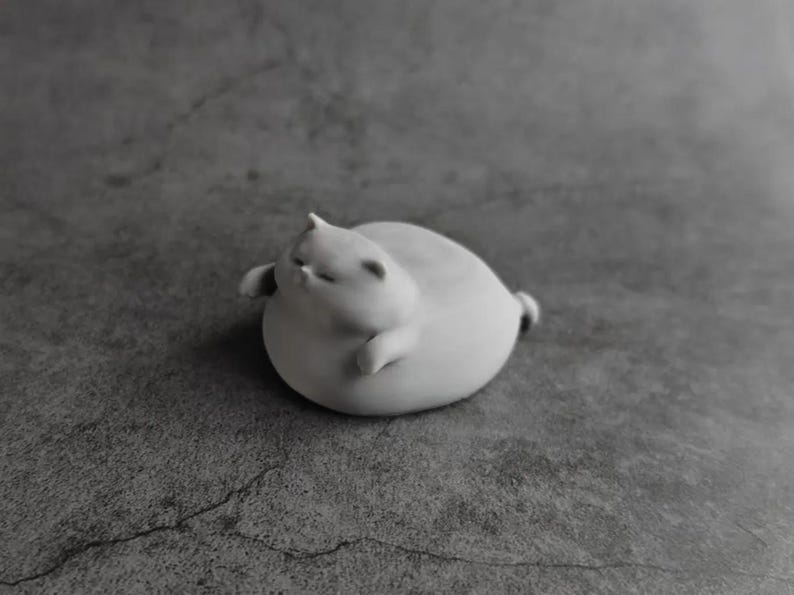May include: A small, white, ceramic cat figurine in a relaxed pose. The cat is round with its eyes closed, small ears, and tiny paws. The figurine is on a gray surface.