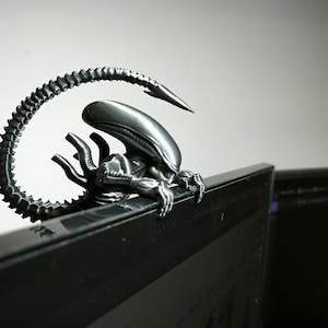 May include: A silver Alien figurine, with a long, curved tail, is perched on a computer monitor. The figurine's design is highly detailed. The background is a plain white wall. The overall aesthetic is sci-fi.