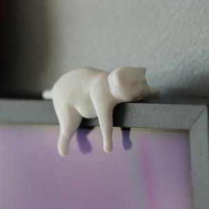 May include: A white ceramic cat ornament, in a relaxed pose, is draped over the edge of a dark grey picture frame. The cat's body hangs down, with its legs dangling. The background is a light grey wall and a purple and white image.