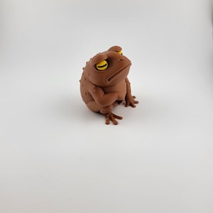 May include: A brown frog figurine with yellow eyes and a grumpy expression. The frog is sitting upright, with its arms crossed and its legs extended. The figurine has a textured surface and is set against a white background.