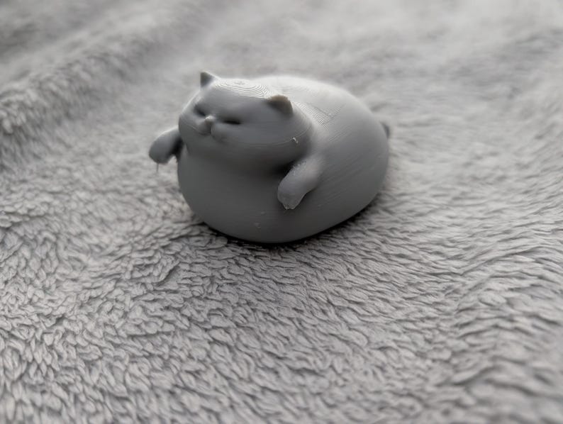 May include: A small, gray, 3D-printed cat figurine. The cat is in a relaxed pose, with its eyes closed and paws extended. The figurine is resting on a soft, gray, textured surface.
