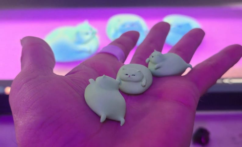 May include: A hand holding three small, light green cat figurines. The cats are in various poses, with one curled up and two standing. The background features blurred, light blue cat shapes.