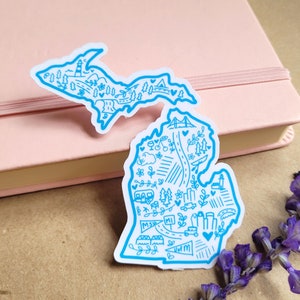 May include: A blue outline sticker of the state of Michigan with various illustrations inside, including trees, a lighthouse, a bridge, a car, and the words "MSU" and "U of M".