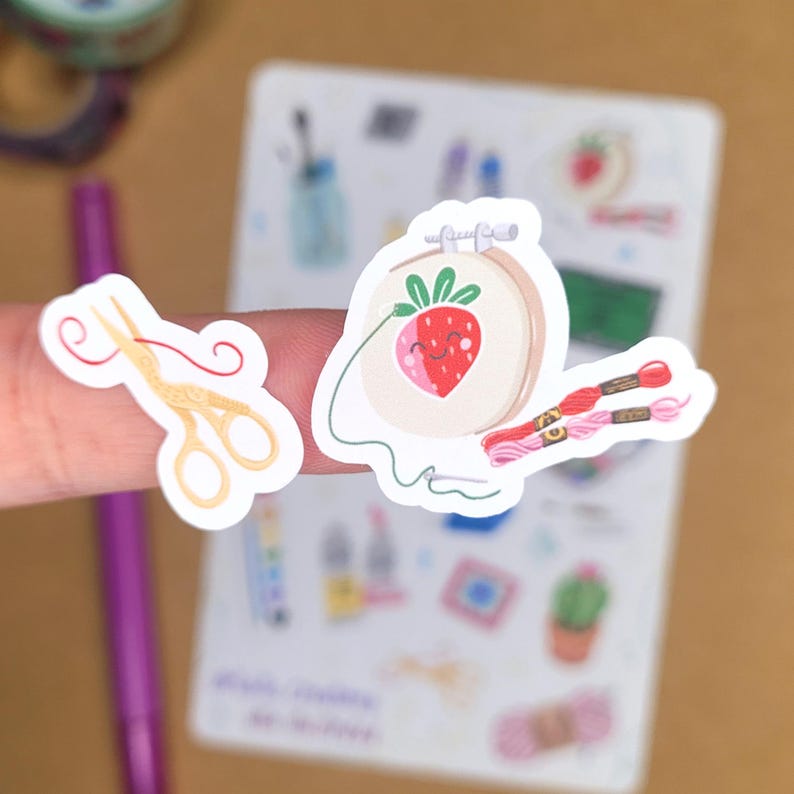 May include: A collection of stickers featuring crafting themes. One sticker shows a smiling strawberry inside an embroidery hoop with thread and floss. Another sticker depicts gold scissors with red thread. The stickers are on a light brown surface.