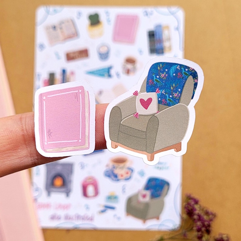 May include: A pink book sticker and a sticker of a gray armchair with a blue floral blanket and a heart-shaped pillow. The stickers are on a background with other small illustrations of cozy items, such as a teacup and a fireplace.