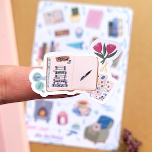 May include: A sticker featuring an open journal with a pen, a stack of books, and the text "Books to read." The sticker also includes a pair of scissors and pink tulips. The background shows other stickers with a cozy theme.