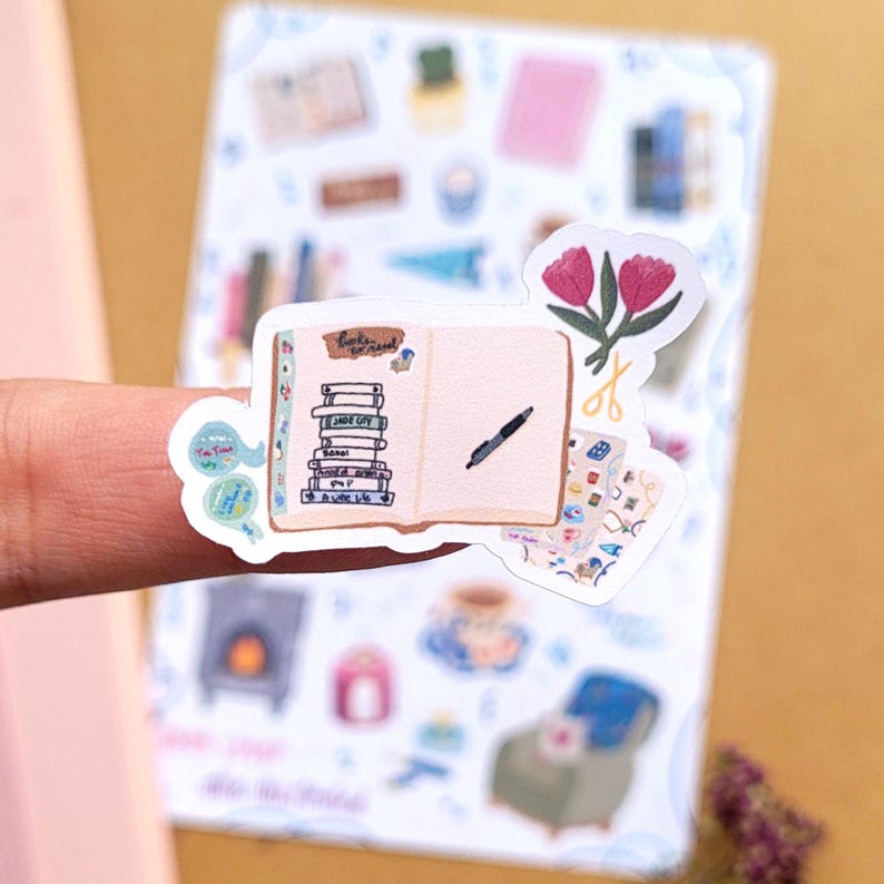 May include: A sticker featuring an open journal with a pen, a stack of books, and the text "Books to read." The sticker also includes a pair of scissors and pink tulips. The background shows other stickers with a cozy theme.