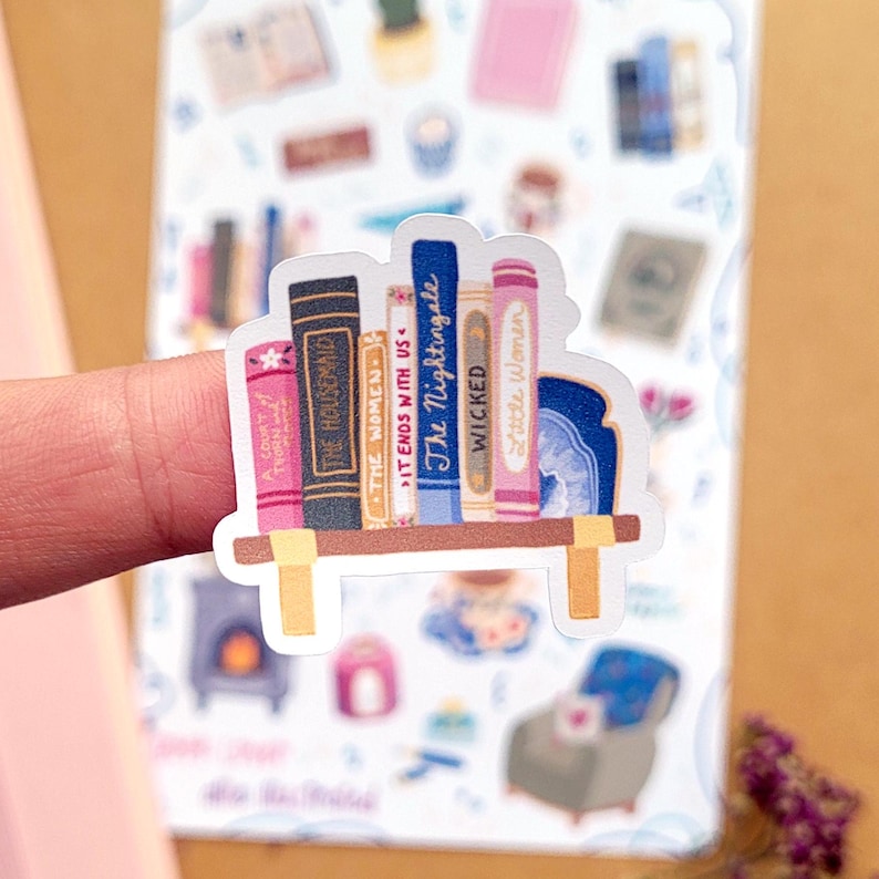 May include: A sticker featuring a bookshelf with colorful book spines. Titles include "The Housemaid," "The Women," "It Ends With Us," "The Nightingale," "Wicked," and "Little Women." The shelf is light brown with yellow supports, and the background includes other stickers.