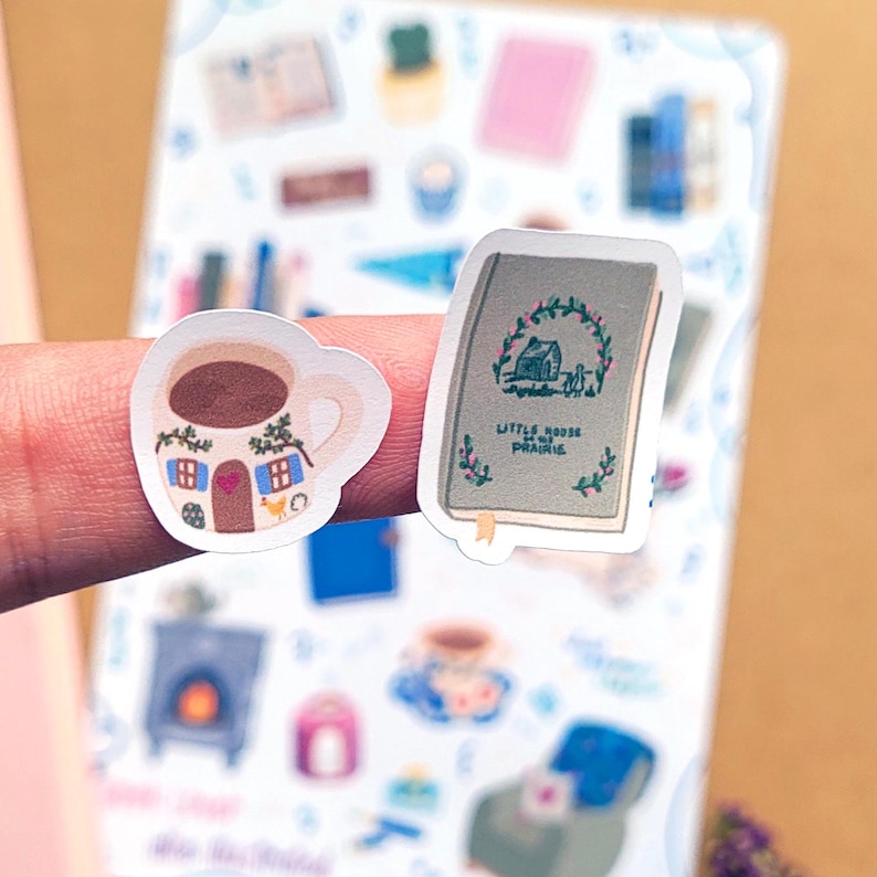 May include: Two stickers: a coffee cup with a house design and a book with the title "Little House on the Prairie". The coffee cup is white with a brown coffee fill, blue windows, and a red heart. The book is green with a house illustration.