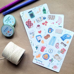May include: Two rolls of washi tape labeled Sunday Garden Club and Cozy Weekend, two purple pens, a spool of white twine, and three sticker sheets featuring illustrations of books, food, drinks, and cozy items with the text Cozy Morning allie illustrated.