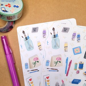 May include: A collection of art supplies and stationery items. Includes stickers with art-themed illustrations, washi tape with the text "COZY WEEKEND", a purple pen, and a variety of stickers featuring paintbrushes, paint tubes, and other art supplies.