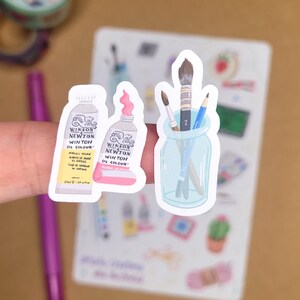 May include: A collection of art-themed stickers. Two stickers depict tubes of Winsor & Newton oil paint, one yellow and one pink. A third sticker shows paintbrushes and pencils in a glass jar. The stickers are on a white background.