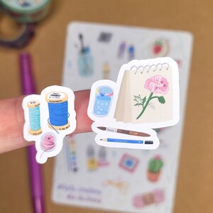 May include: A collection of stickers featuring art and craft supplies. The stickers include spools of thread, a notepad with a pink flower, a glass of water, a paintbrush, and a pencil. The stickers are colorful and have a hand-drawn aesthetic.